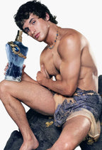 A shirtless man with dark curly hair sits on a dark rock against a white background, holding a blue perfume bottle with gold detailing. He is wearing patterned blue and beige shorts and a beaded necklace.