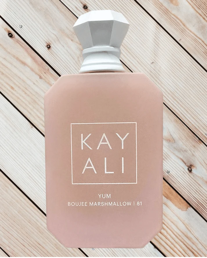 A dusty pink bottle of Kayali Yum Boujee Marshmallow | 81 with a white faceted cap, displayed on a light wood plank background.